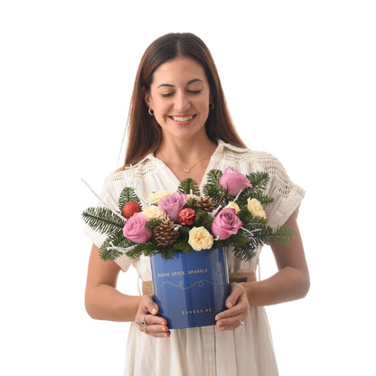 Woman smiles holding festive blue floral arrangement with pink roses, pine, and ornaments.