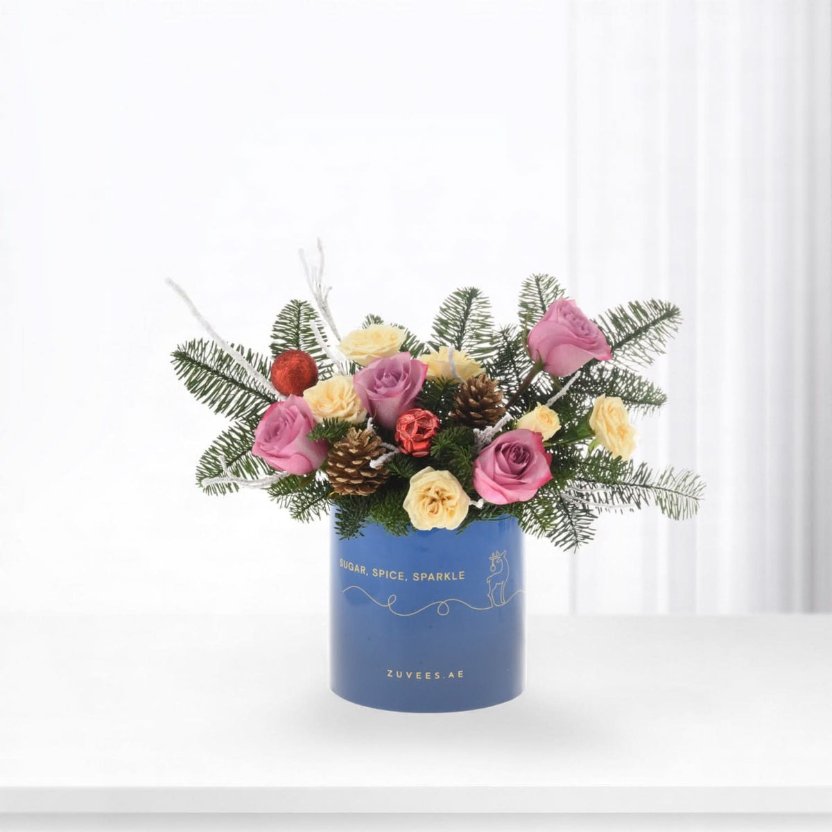 Blue vase with pink, cream roses, pine, ornaments, "Sugar, Spice, Sparkle" text, festive holiday bouquet.