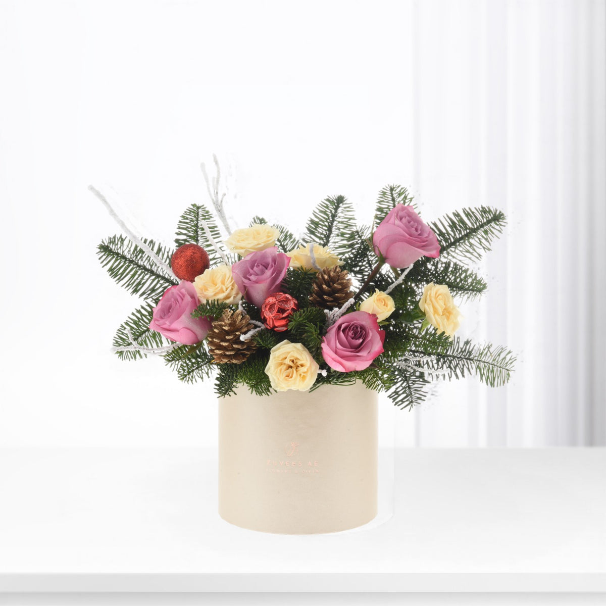 Pink and cream roses with pinecones, red ornaments, and evergreen sprigs in a beige cylinder vase.