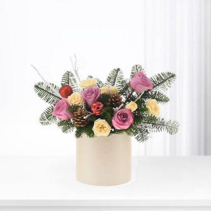 Pink and cream roses with pinecones, red ornaments, and evergreen sprigs in a beige cylinder vase.