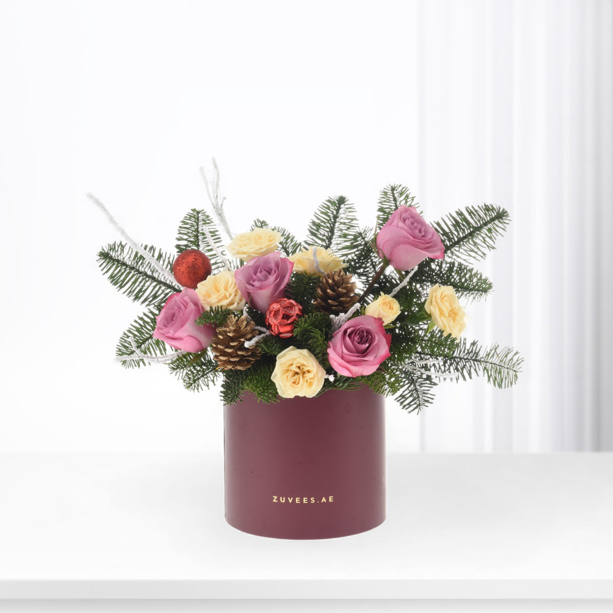 Pink and yellow roses nestled in evergreens with pinecones and red ornaments in burgundy cylinder.