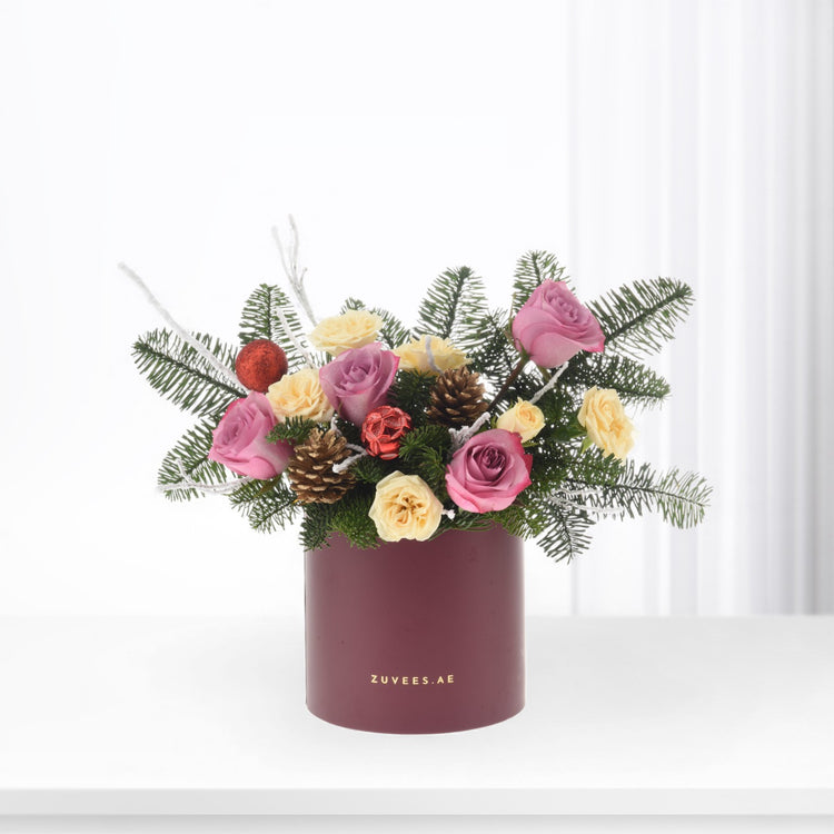 Pink and yellow roses nestled in evergreens with pinecones and red ornaments in burgundy cylinder.