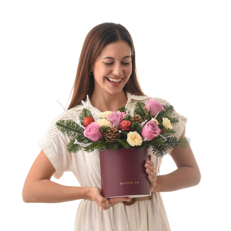 Woman smiles holding burgundy box with pink, yellow roses, pine, and ornaments.