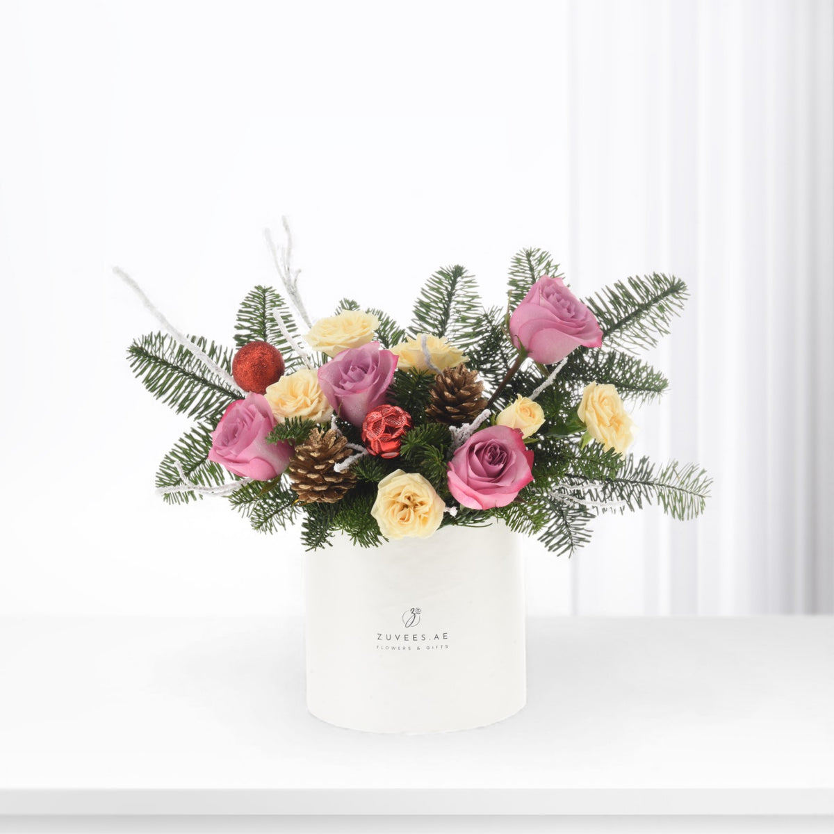 White vase holds pink, yellow roses, pine, pinecones, and red ornaments.