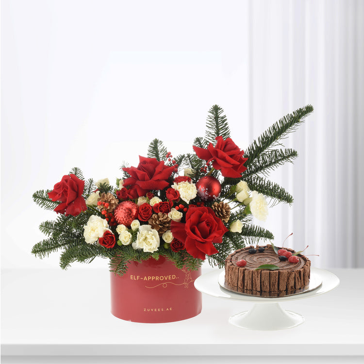 Vibrant red roses, pine, and ornaments in festive red box beside chocolate cake.