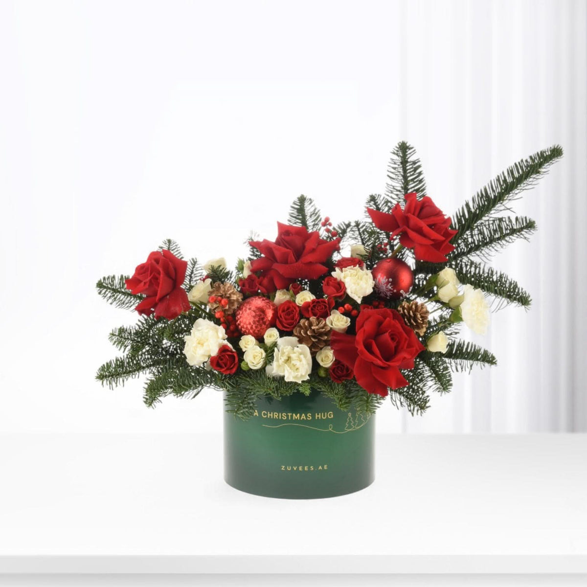 Vibrant red roses, white blooms, pine, and ornaments in festive green box.