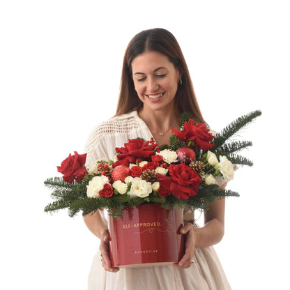 Woman holds festive red-and-white floral arrangement in glossy "Elf-Approved" box.