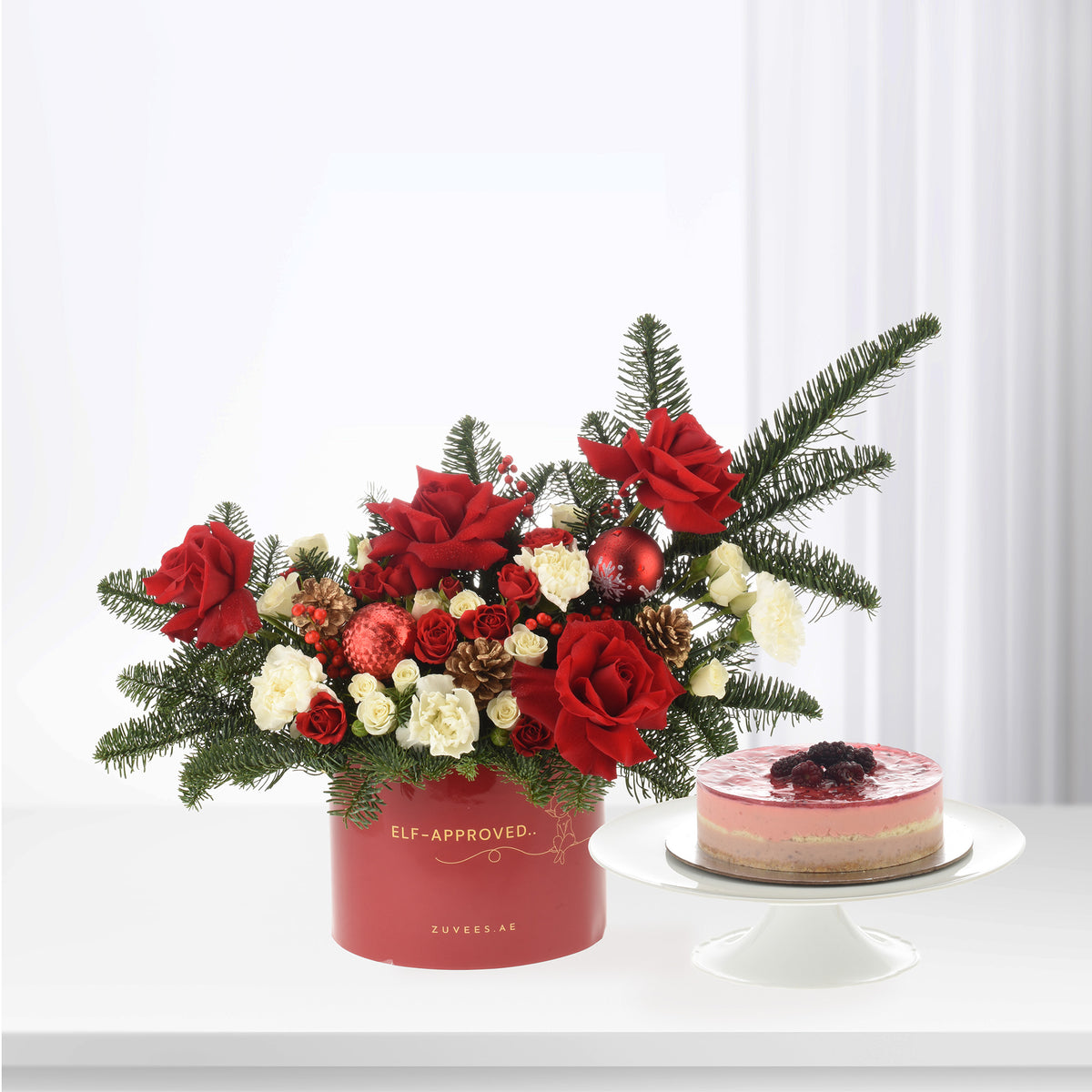 Festive red roses, pine, ornaments in "Elf-Approved" box beside berry-topped cheesecake on white stand.