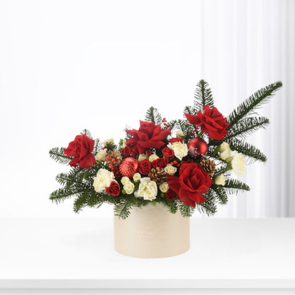 Vibrant red roses, white carnations, pine, and ornaments in elegant beige box.