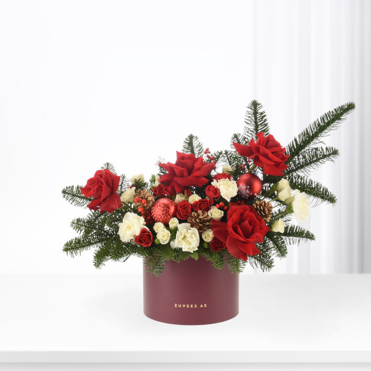 Vibrant red roses, white blooms, pine, pinecones, and ornaments in burgundy box.