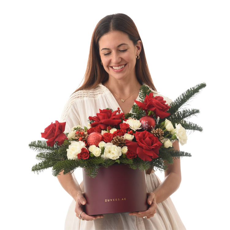 Woman holds festive red and white rose bouquet with pine, ornaments in burgundy Zuvees box.