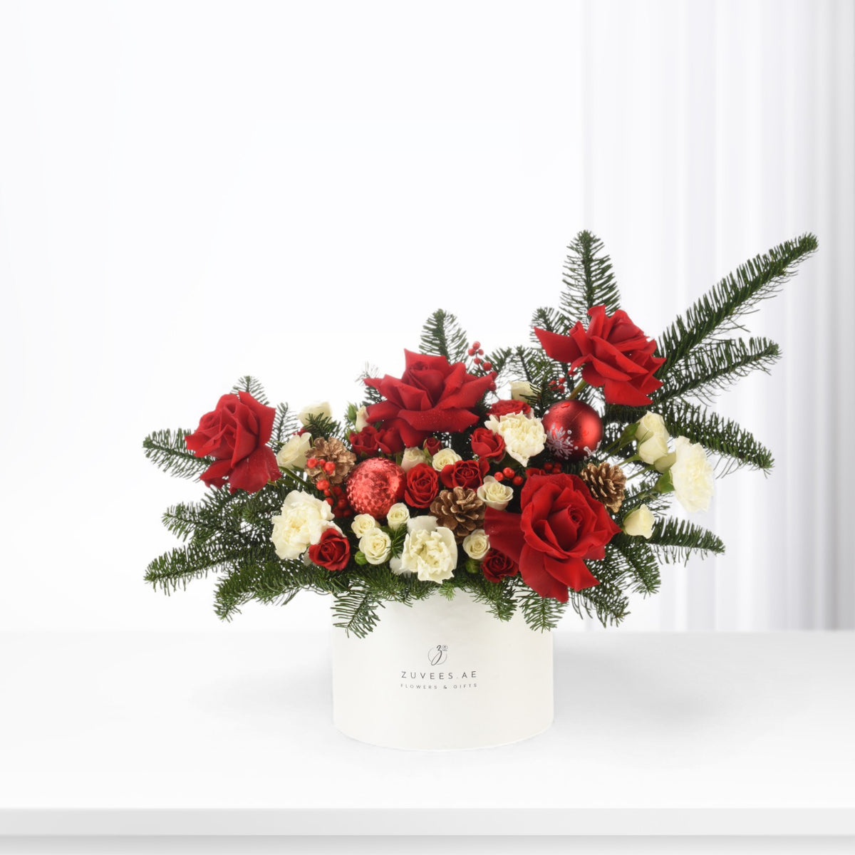 Vibrant red roses, white carnations, pine, pinecones, and red ornaments in white box.