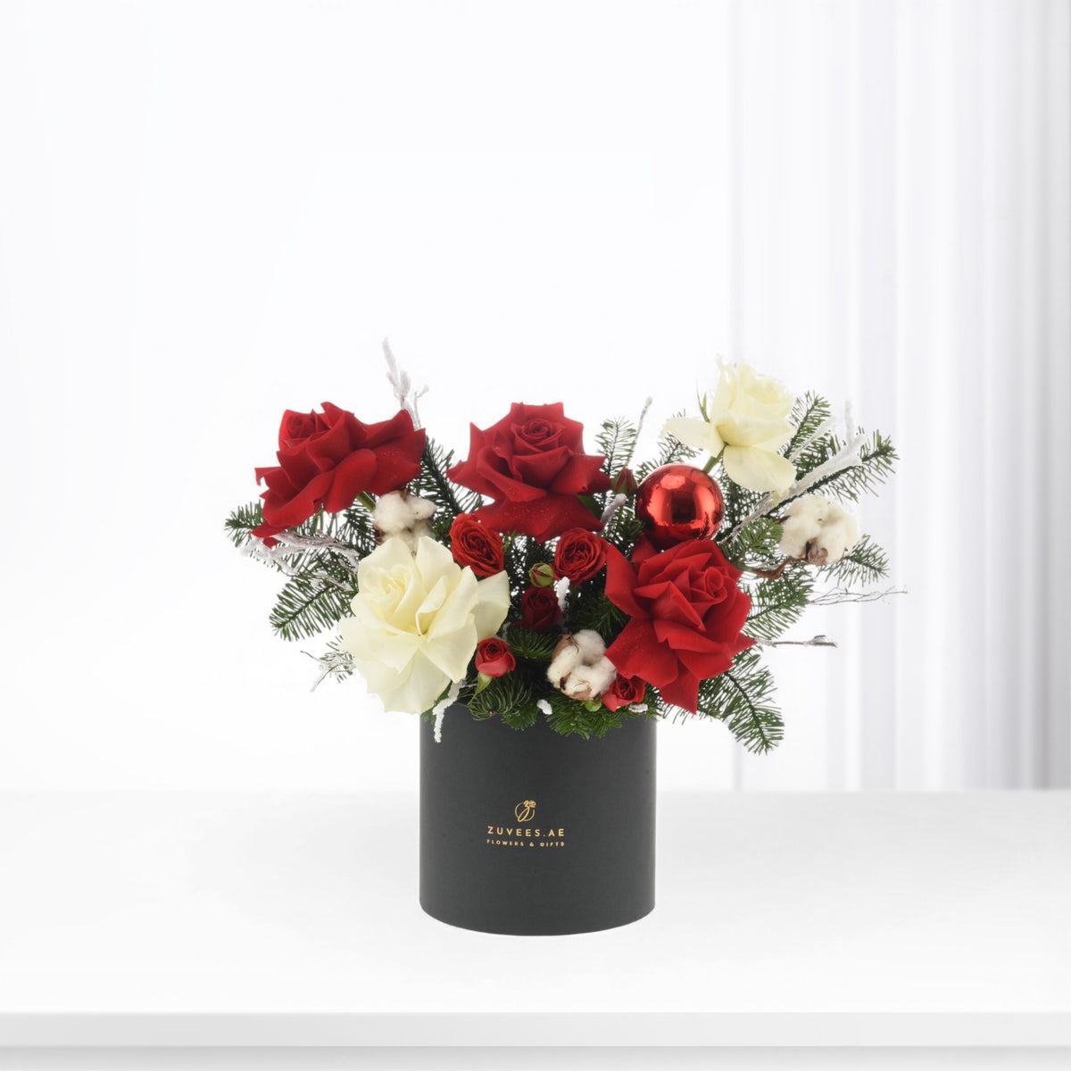 Elegant black vase holds vibrant red and cream roses, pine, cotton, and a shiny red ornament.