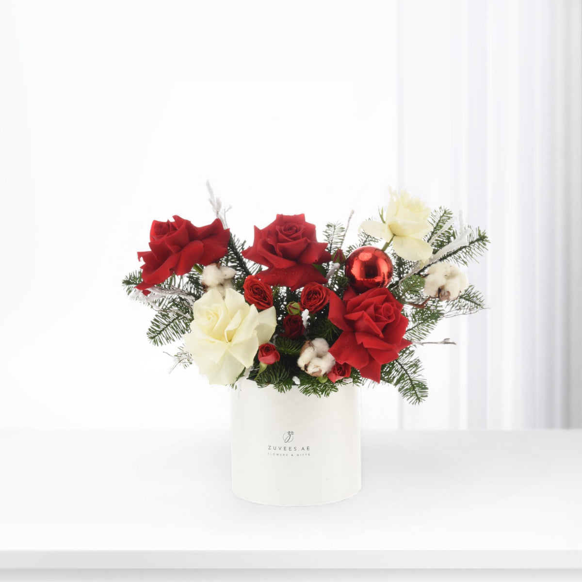 Vibrant red and white roses, cotton, pine, and a red ornament in a branded white vase.