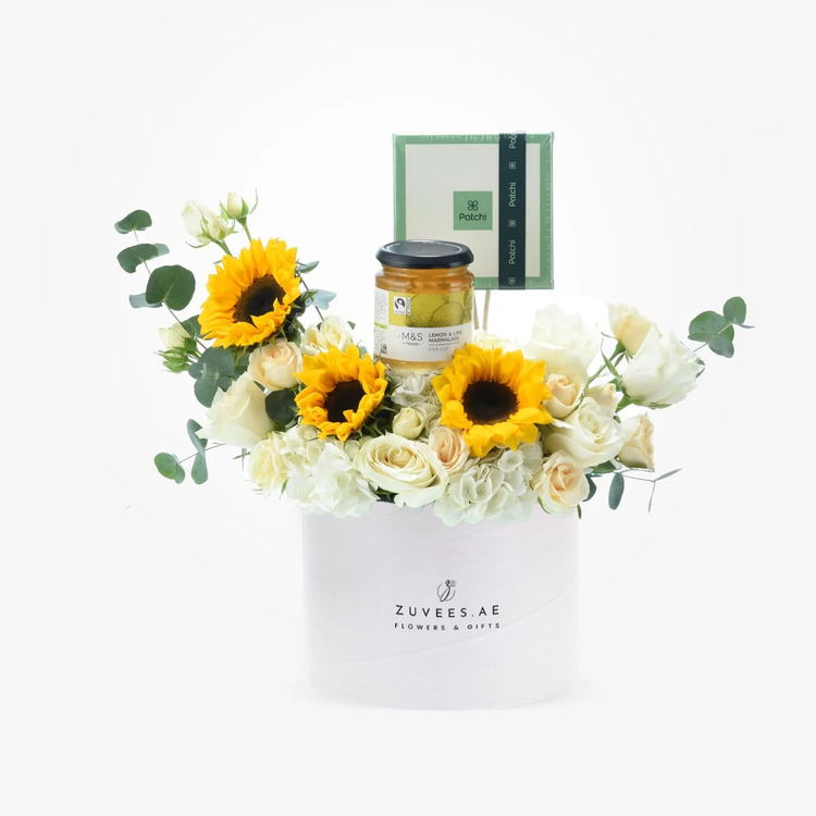 White cylinder holds vibrant sunflowers, creamy roses, and gourmet gifts.