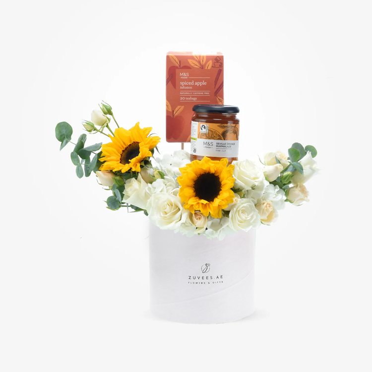 White cylindrical box holds vibrant sunflowers, white roses, and greenery, topped with M&amp;S tea and marmalade.