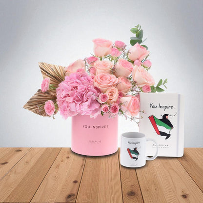 Pink roses and hydrangeas in branded pink vase, matching mug and notebook on wooden table.