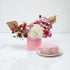 Pink floral arrangement with gold accents beside matching cake on marble.