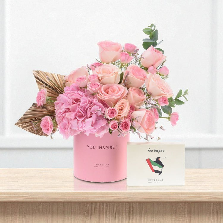 Pink roses and hydrangeas in a "You Inspire!" cylinder, beside matching card on wood.