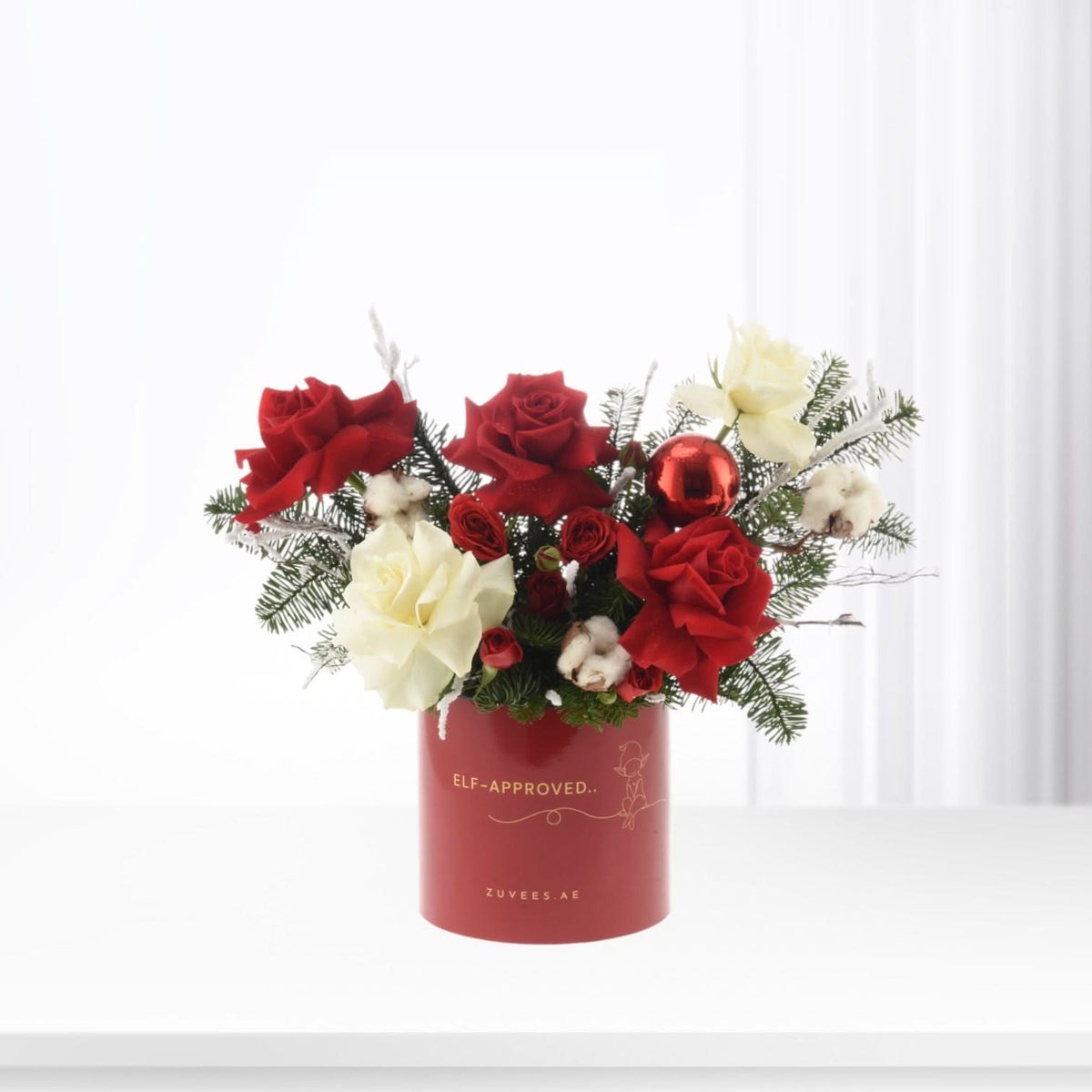 Vibrant red and white roses, pine, cotton, and ornament in festive red cylindrical vase.