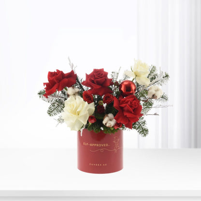 Vibrant red and white roses, pine, cotton, and ornament in festive red cylindrical vase.
