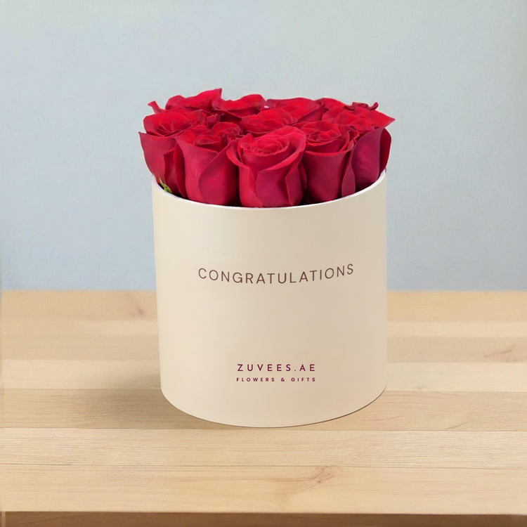 Vibrant red roses in cream "Congratulations" box on wooden surface.
