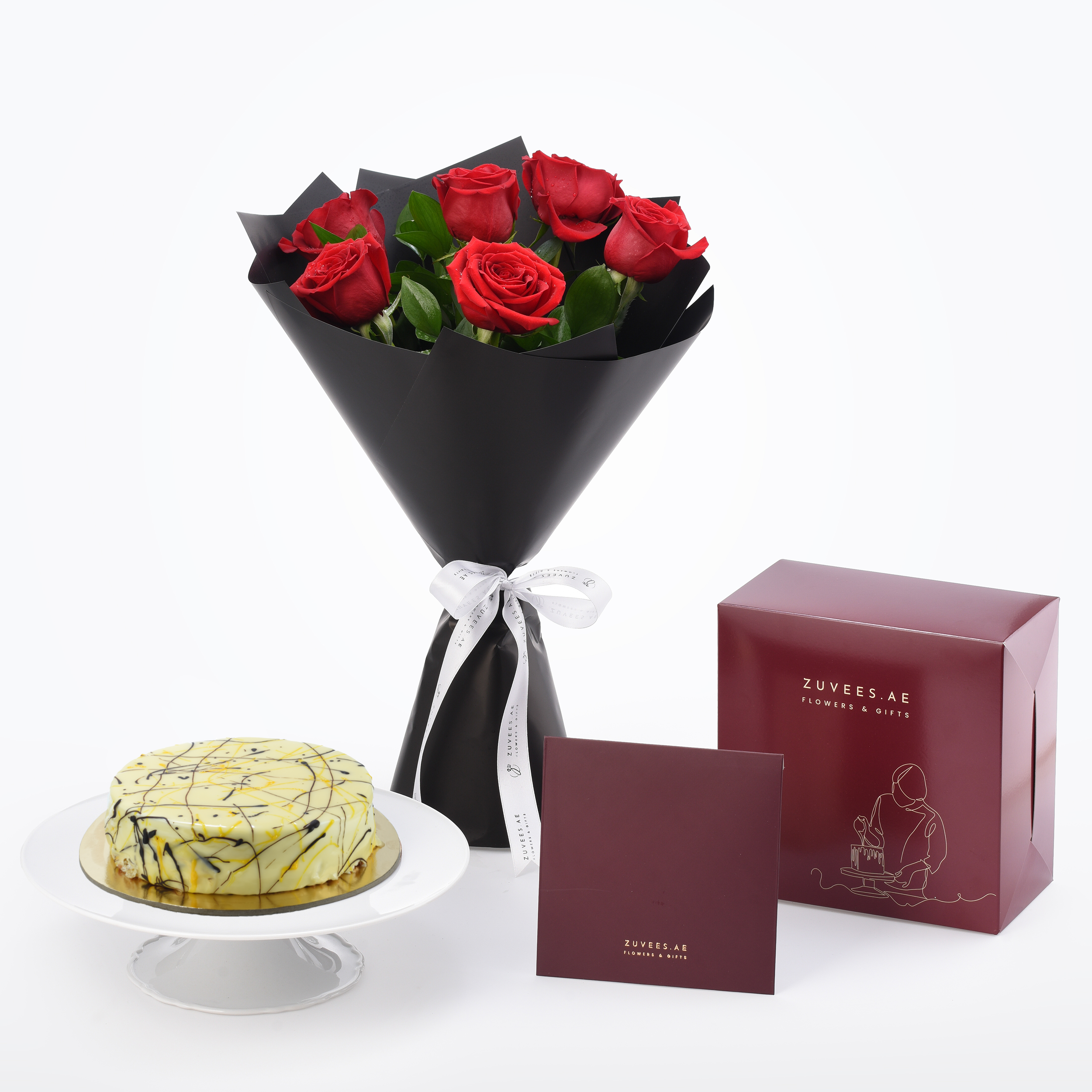 Vibrant red roses in black wrap beside marble cake and burgundy gift box.