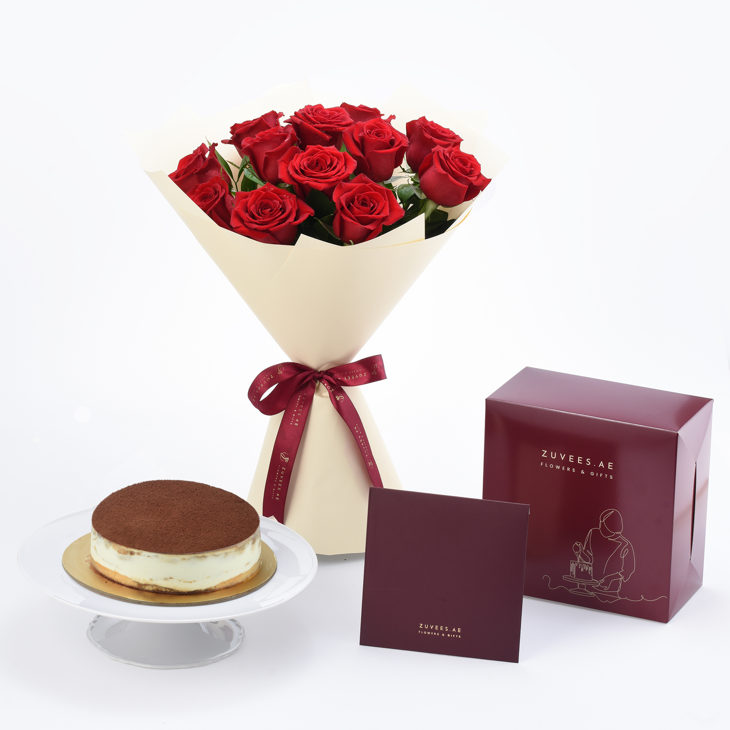 Vibrant red roses in cream wrap with burgundy ribbon, beside chocolate cake and branded gift box.