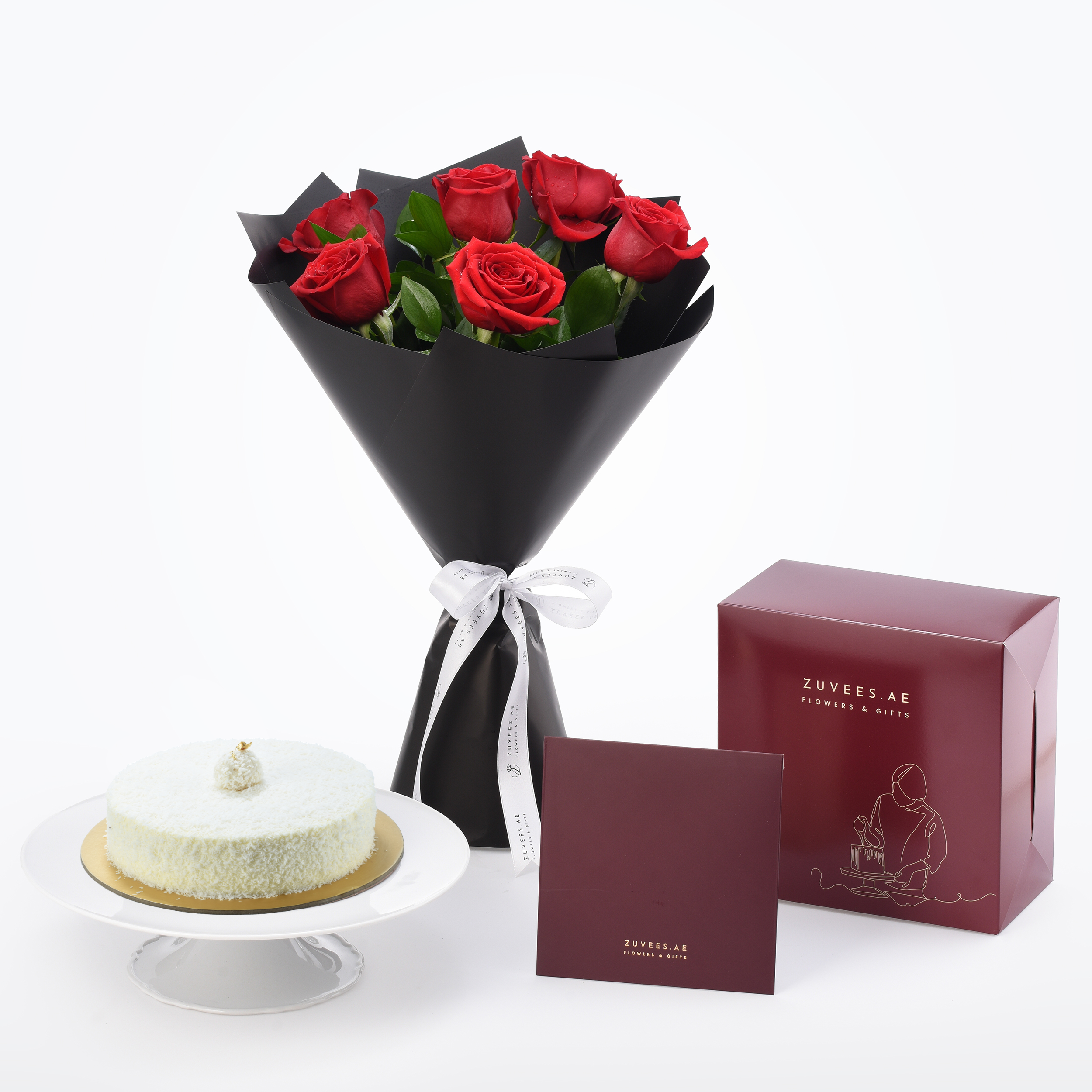 Vibrant red roses in black wrap beside coconut cake and burgundy gift box.