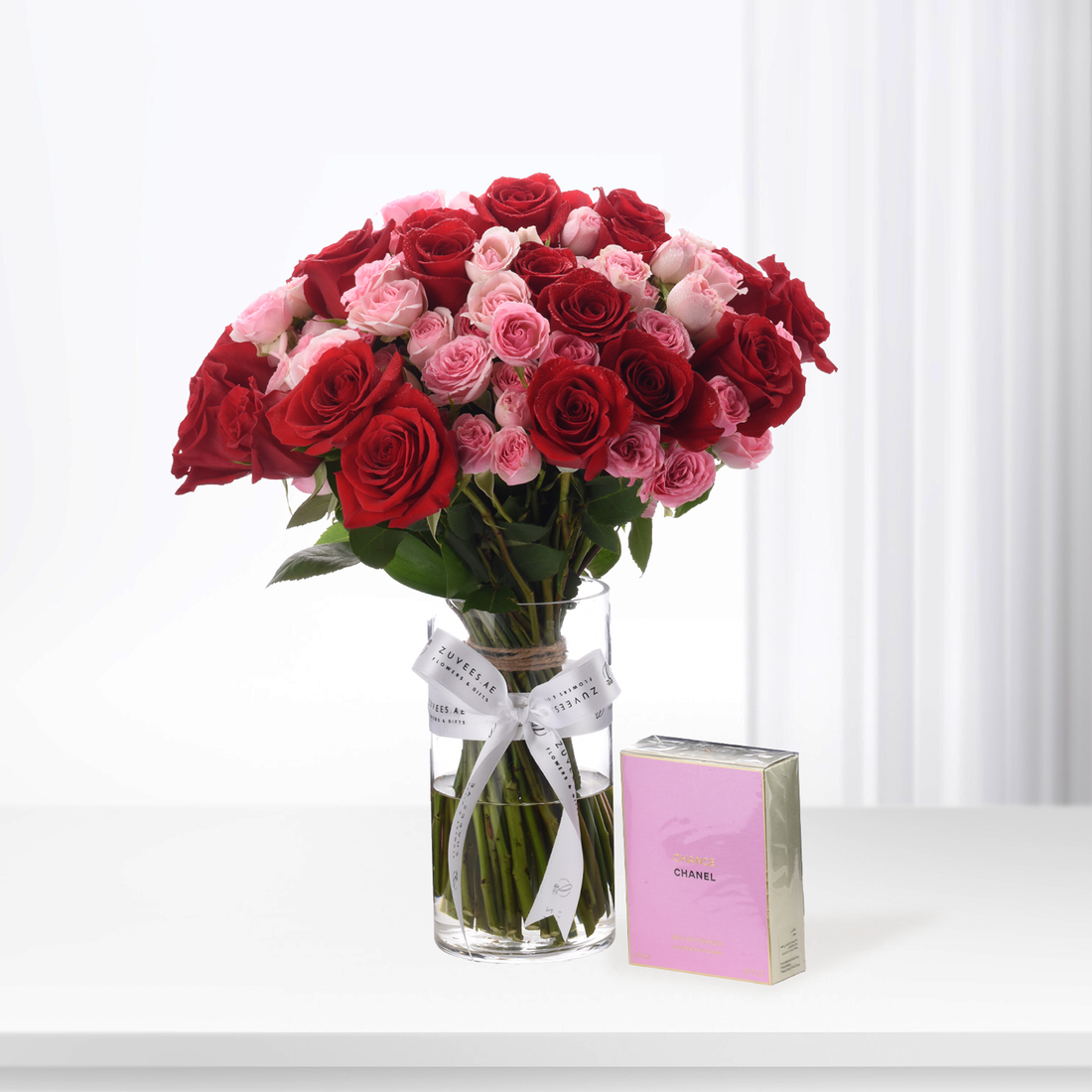 Vibrant red and pink roses in glass vase with white ribbon, beside pink Chanel perfume box.