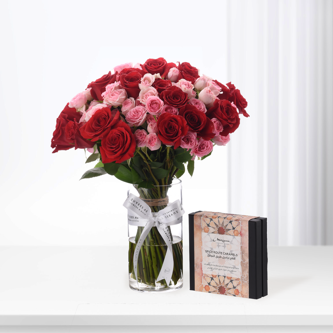 Vibrant red and pink roses in glass vase beside ornate caramel gift box.