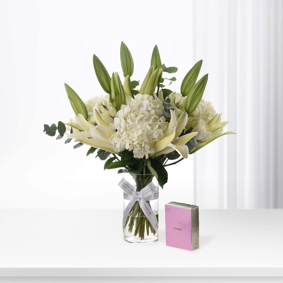 White hydrangeas, lilies, eucalyptus in clear vase with ribbon, beside pink Chanel box on white surface.
