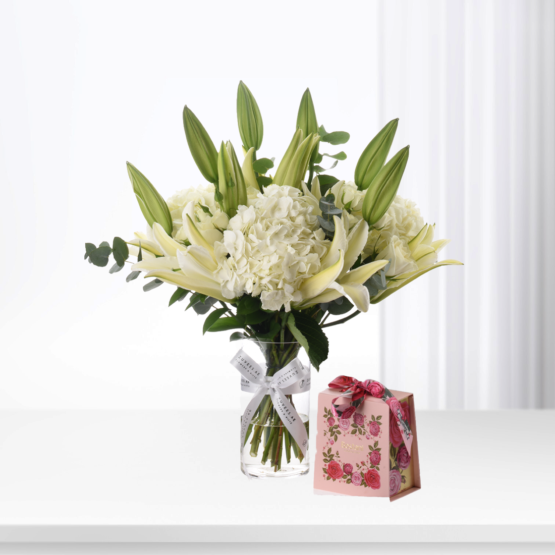 Elegant white lilies, hydrangeas, and buds in glass vase beside pink rose gift box.