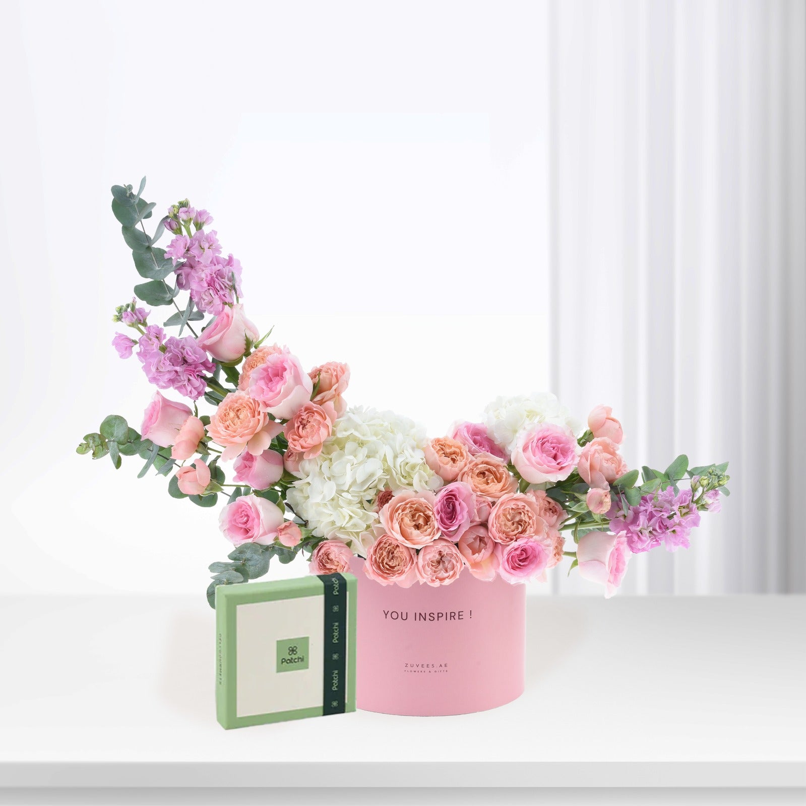 Pink rose bouquet with hydrangeas, eucalyptus, in pink "YOU INSPIRE!" box beside green Patchi chocolate.