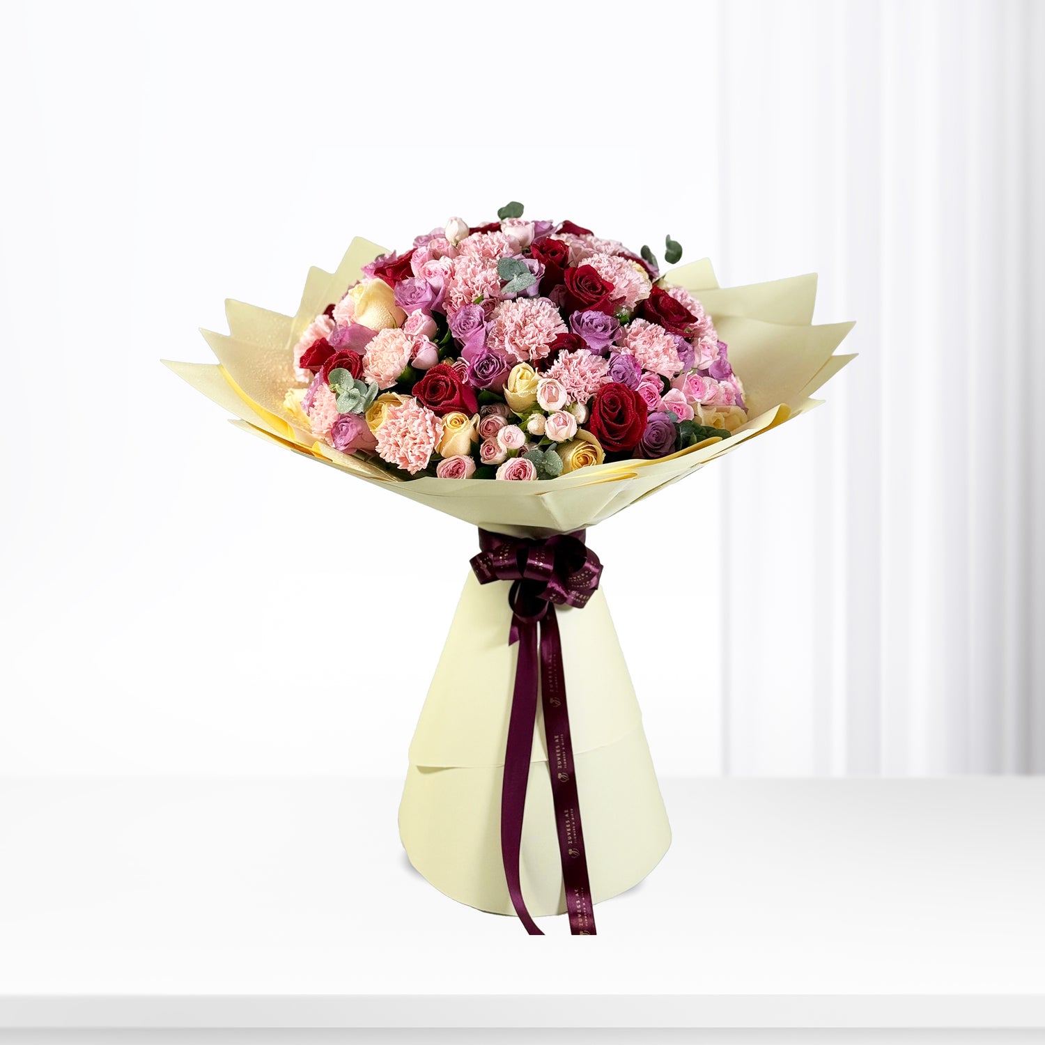 Vibrant pink, red, and yellow roses and carnations wrapped in cream paper with burgundy ribbon.