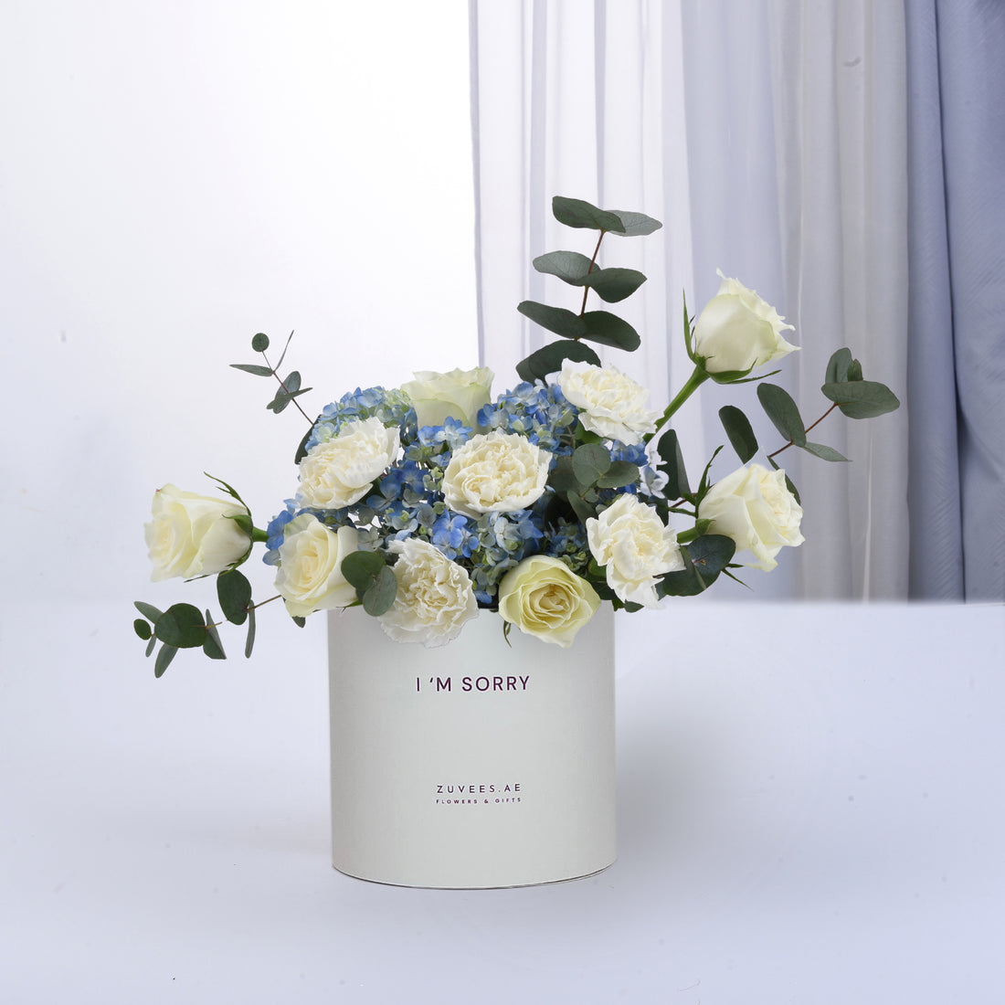 White roses and blue hydrangeas in minimalist white box with "I&