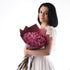 Woman in white lace dress holds burgundy-wrapped bouquet of pink roses against clean white background.