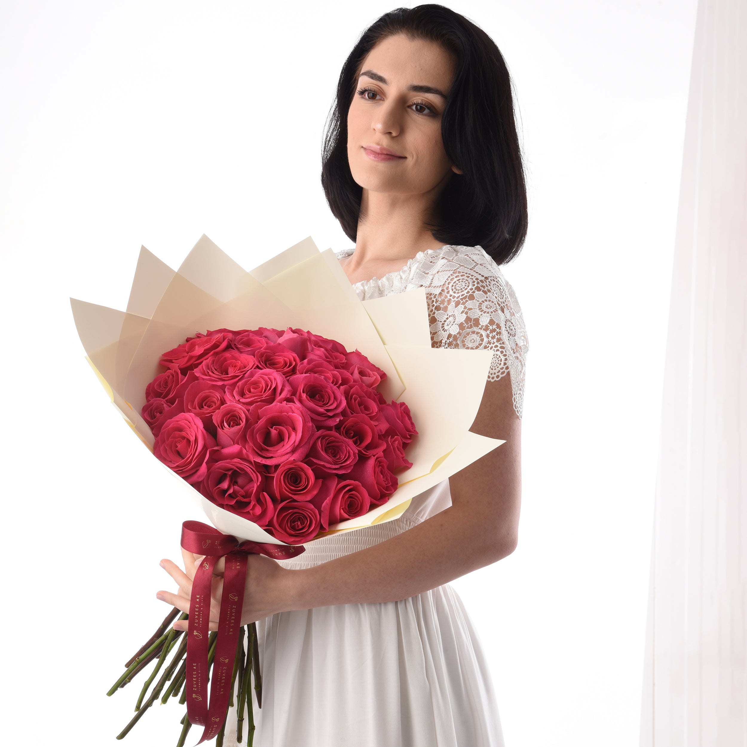Woman holds vibrant red rose bouquet wrapped in cream paper with burgundy ribbon.