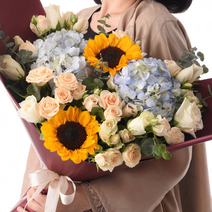 Vibrant sunflowers, blush roses, and blue hydrangeas in burgundy wrap.