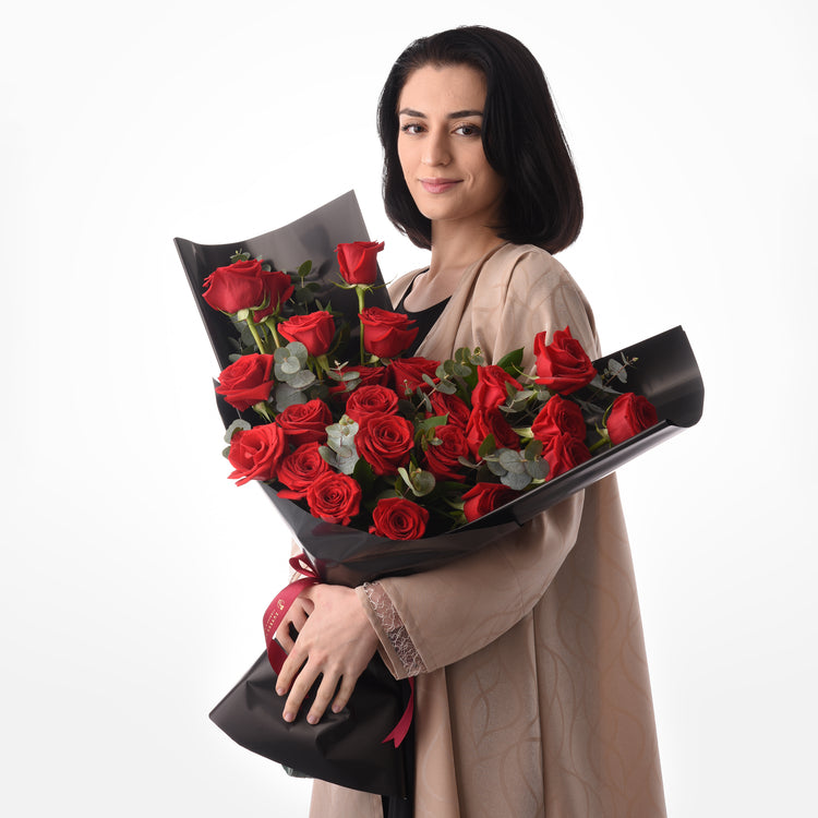 Woman holds lush red rose bouquet in sleek black wrap against white background.