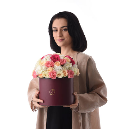 Woman holds burgundy box with vibrant pink, peach, white roses and carnations, gold logo.
