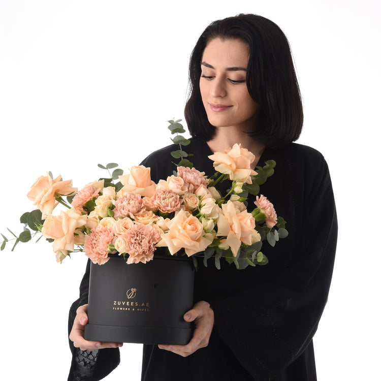 Elegant peach roses and carnations in sleek black Zuvees box, held by woman in black attire.