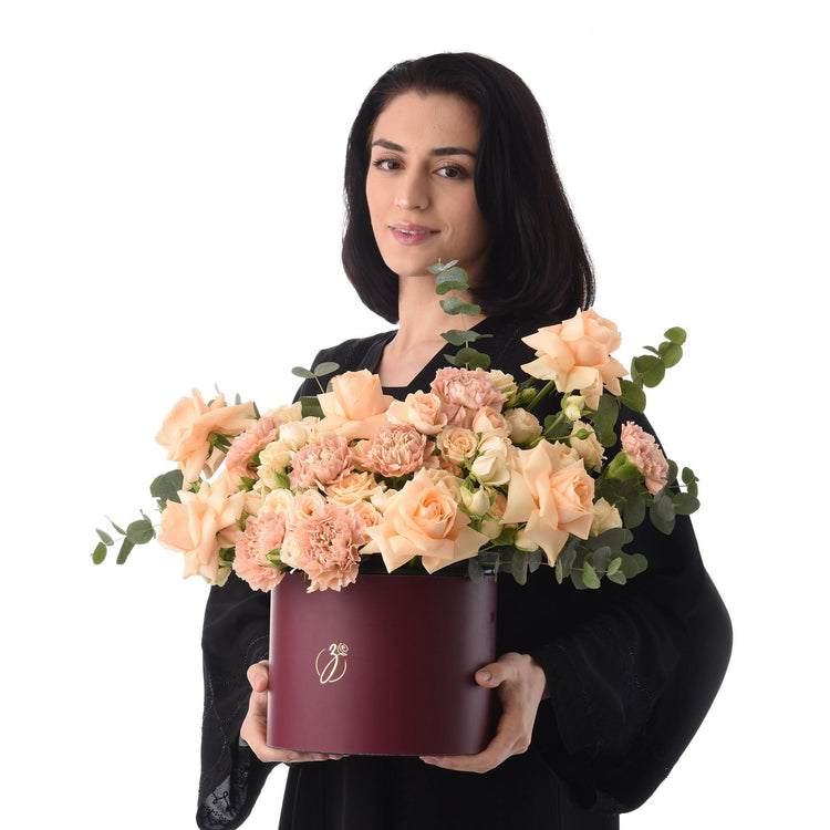 Woman holds burgundy hat box overflowing with peach roses and eucalyptus.