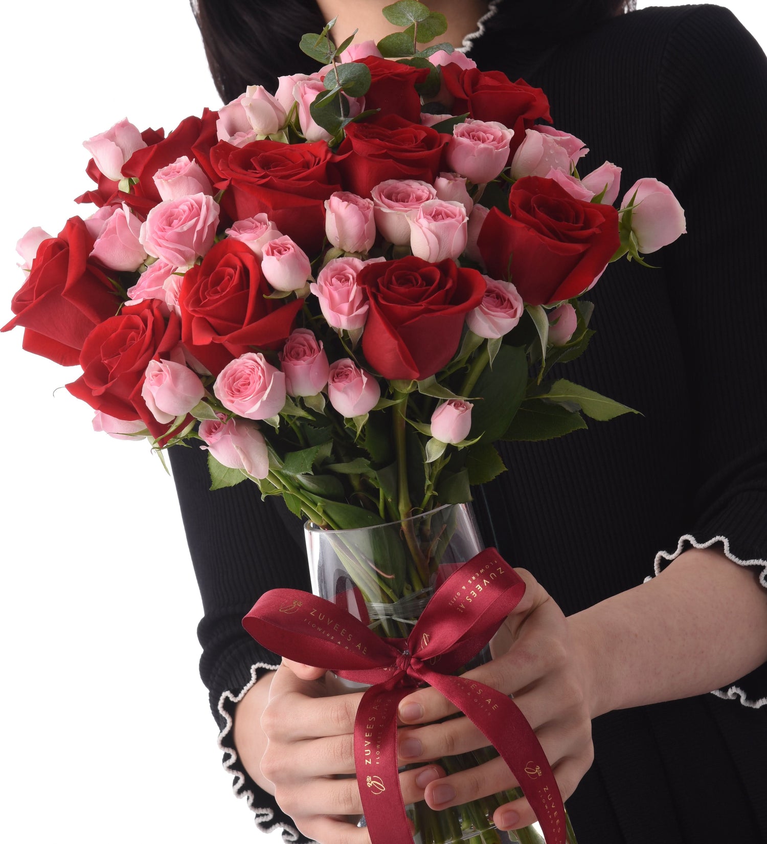 Vibrant red and pink roses in clear vase, tied with branded burgundy ribbon, held by person in black.