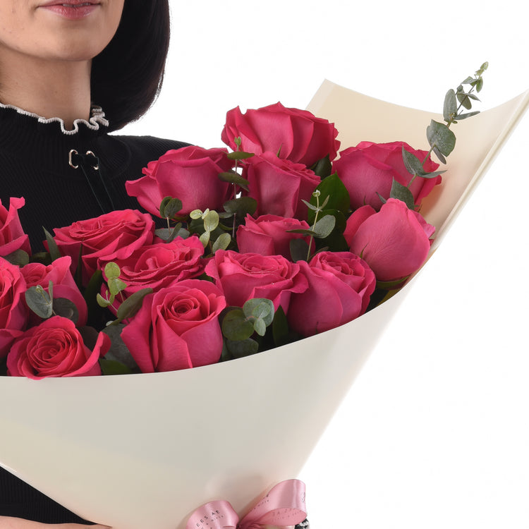 Vibrant pink roses wrapped in cream paper with pink bow, held by woman in black top.