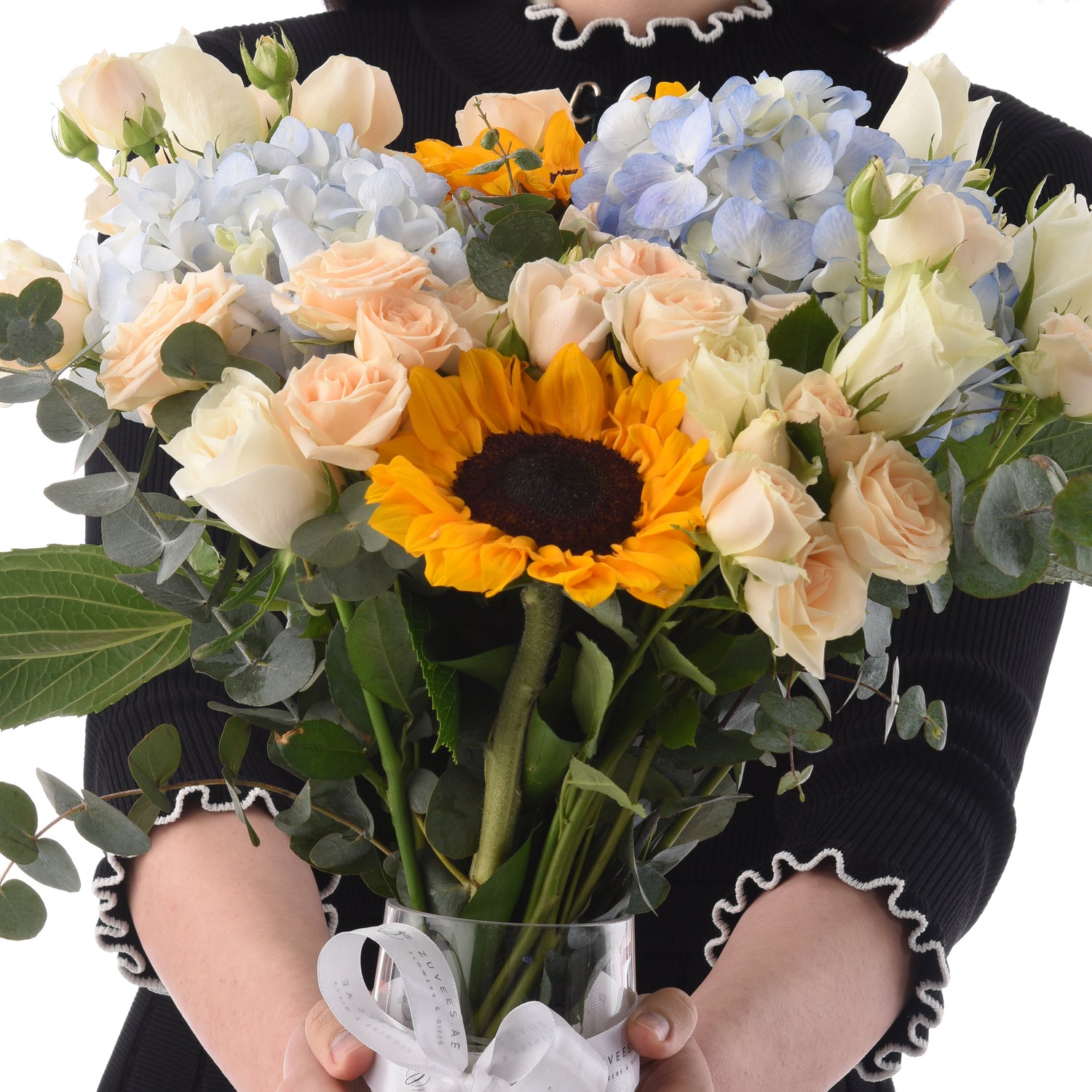 Vibrant sunflower, peach roses, blue hydrangeas, eucalyptus in clear vase, held by person in black.