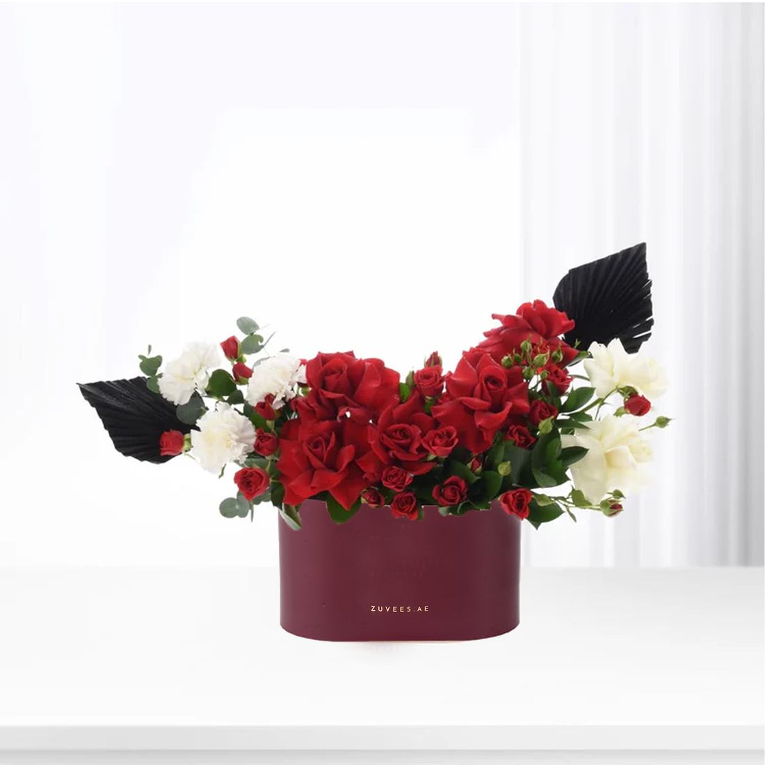 Bold red roses contrast white blooms and black fan leaves in deep burgundy box.