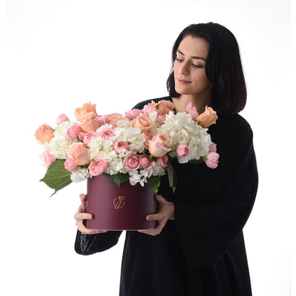 Woman holds burgundy hat box overflowing with soft pink roses and white hydrangeas.