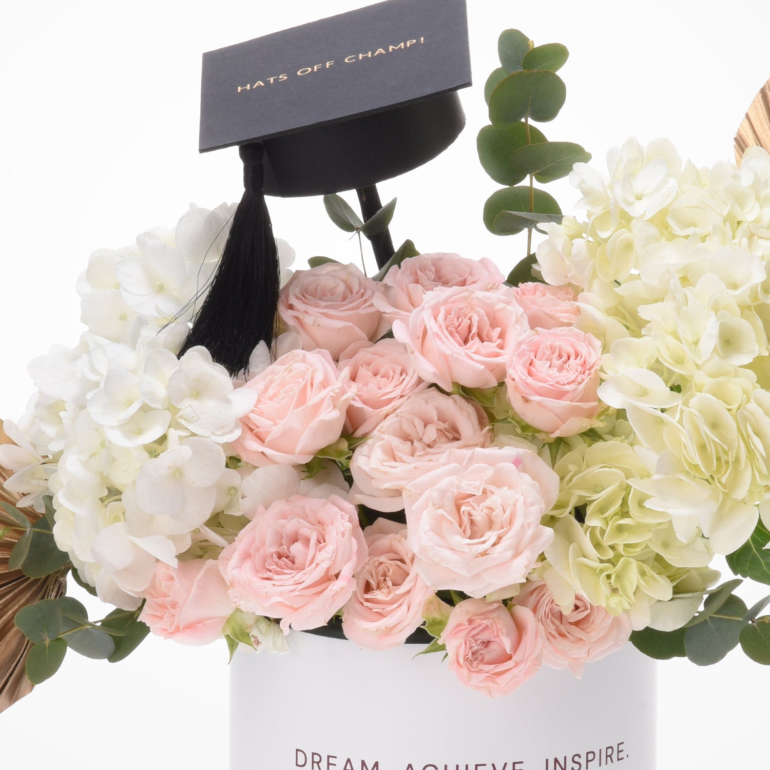 Soft pink roses, white hydrangeas, and eucalyptus surround a graduation cap with “Hats Off Champ!” tag.