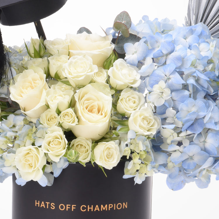 Elegant white roses and blue hydrangeas in black "Hats Off Champion" box.